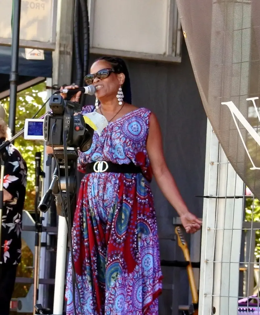 Nadine States performing on an outdoor festival stage in daylight, singing into a microphone in a patterned red and blue dress, one arm outstretched